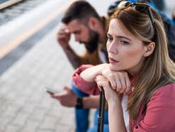 Zwei Menschen auf dem Bahnsteig schauen unzufrieden und haben Koffer dabei.