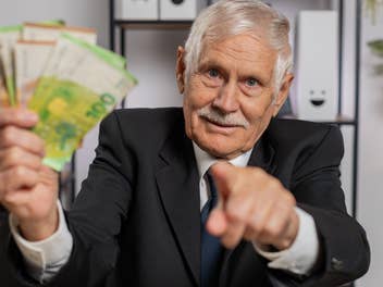 Ein älterer Mann im Anzug hält Geldscheine in einer Hand und zeigt mit dem Finger ins Bild