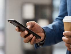 Mann hält Handy in der einen und To-Go-Kaffee in der anderen Hand.