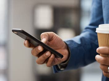 Mann hält Handy in der einen und To-Go-Kaffee in der anderen Hand.