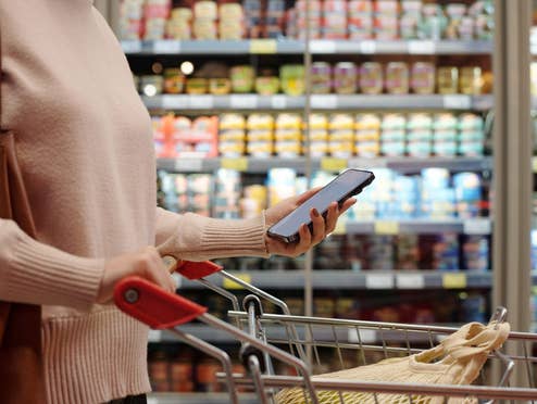 Eine Frau geht mit Einkaufswagen und ihrem Handy durch den Supermarkt.