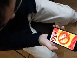 Jugendlicher hält sein Smartphone in der Hand, darauf sind ein Stopp-Zeichen und der Hinweis +16 zu sehen