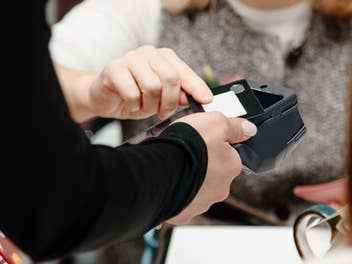 Eine Person hält seine Bankkarte an ein Kartenlesegerät, dabei greift die Kassiererin nach der Karte.