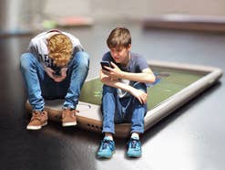 Zwei Kinder nutzen Smartphones, dabei sitzen sie symbilisch auf einem übergroßen Smartphone