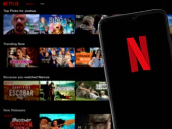 Smartphone mit Netflix-Logo dahinter ein Blick auf verschiedene Serien und Filme bei Netflix