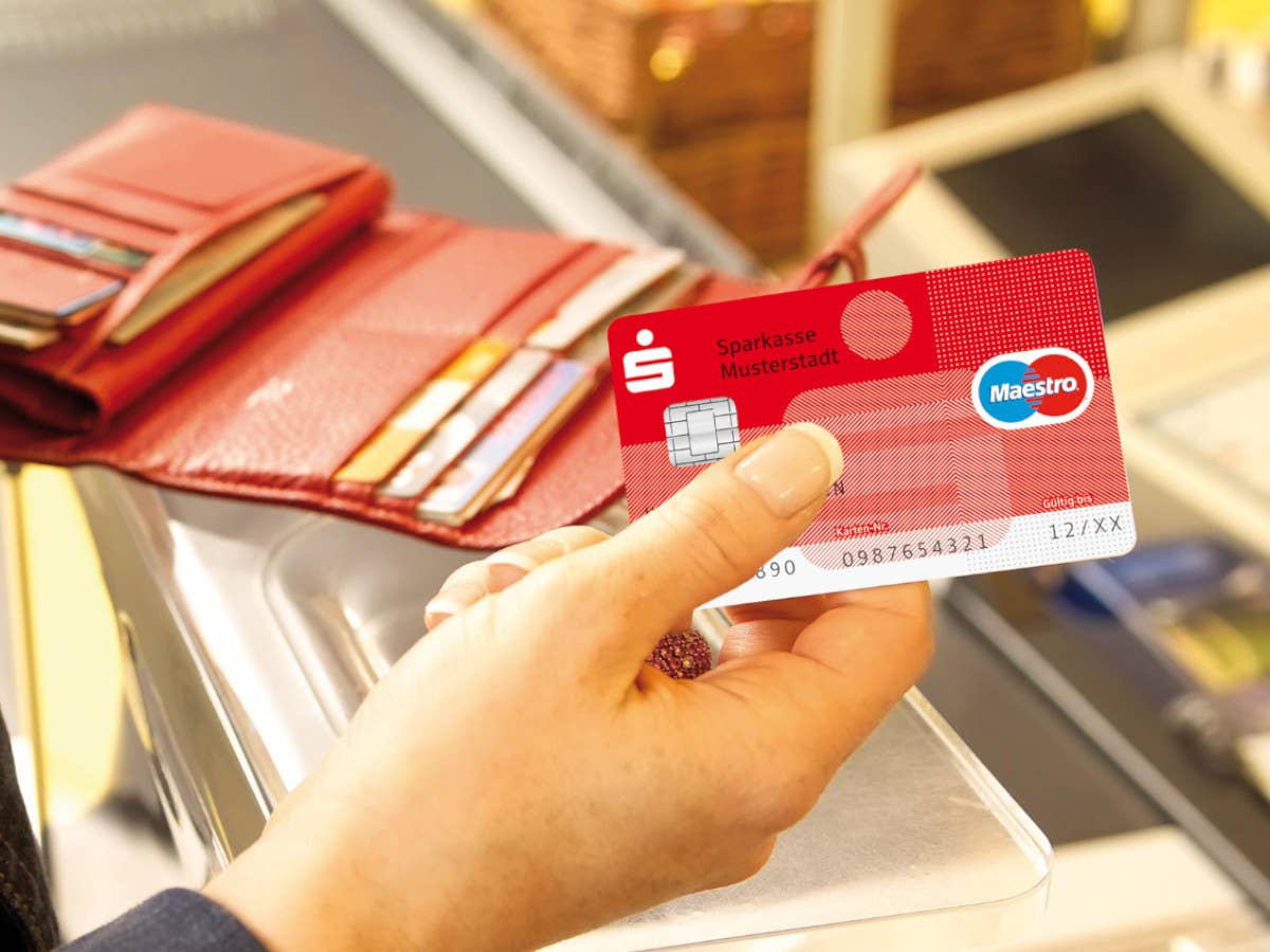 Eine Debitkarte in einer Hand, im Hintergrund ein Portmonnaie und eine Kasse.