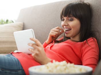Frau isst Popcorn auf dem Sofa und hält dabei ein Tablet PC in der Hand.