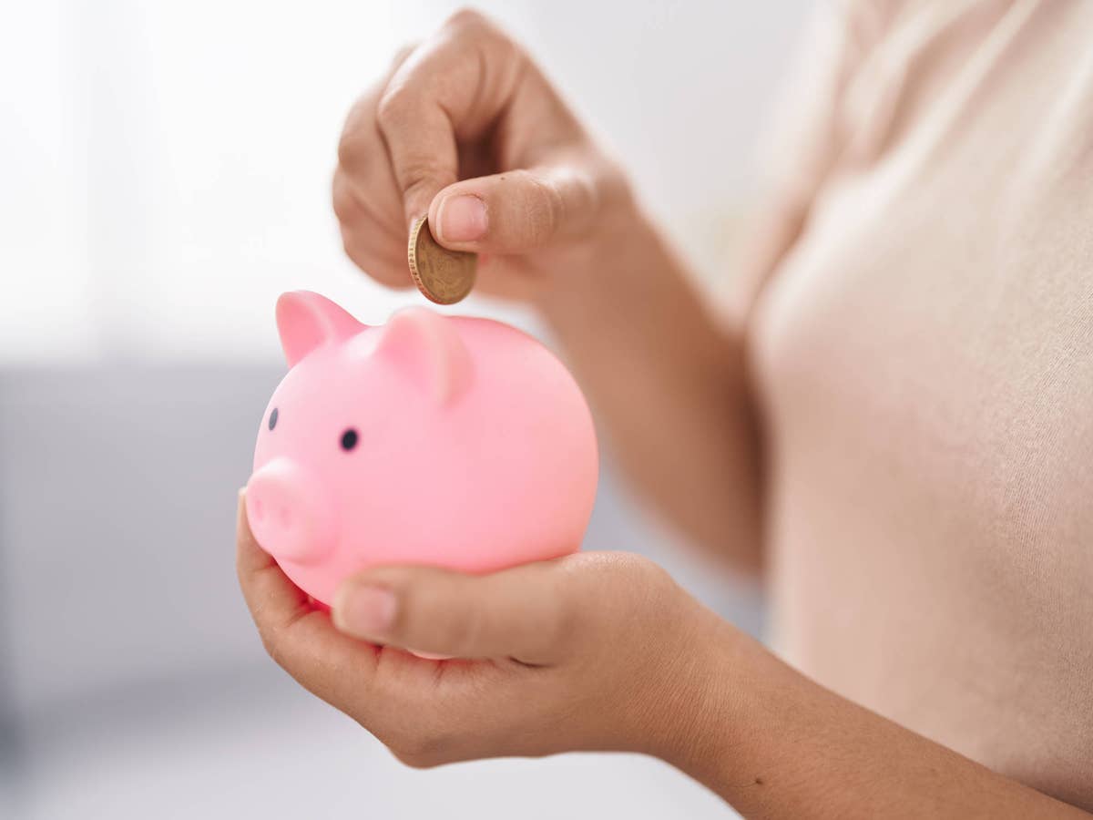 Frau steckt eine Münze in ein Sparschwein, das auf ihrer linken Hand steht.