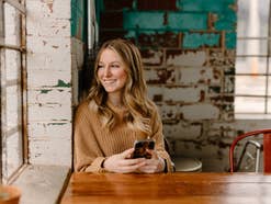 Frau sitzt mit einem Smartphone in der Hand an einem Holztisch und lächelt in Richtung eines Fensters.