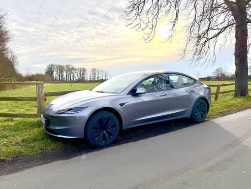 Tesla Model 3 im Sonnenlicht an einem Feldweg unter einem Baum geparkt.