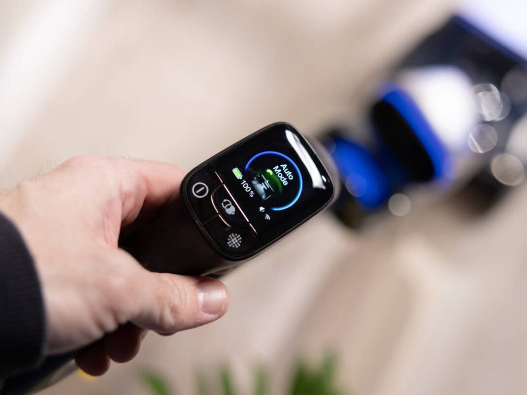 Eine Hand hält den Griff des Staubsaugers, auf dessen kreisförmigem Display "Auto Mode" und ein Akkustand von 100% zu sehen sind.