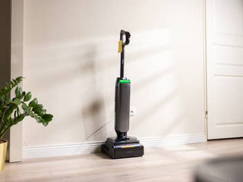 Ein moderner, dunkler Akku-Nass-Trockensauger steht aufrecht an einer weißen Wand auf hellem Holzboden.