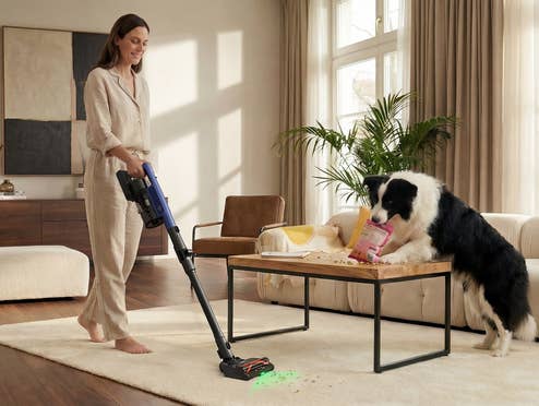 eine Frau saugt ihr Wohnzimmer während ein Hund Trockenfutter vom Couchtisch auf den Boden verstreut