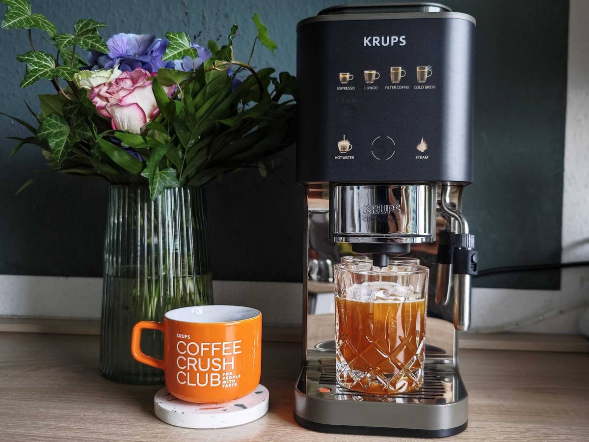 Die Krups Coffee Crush kann viel: Espresso, Cold Brew, Filterkaffee – aber schmeckt der auch gut? Krups Coffee Crush Experience Espresso-Vollautomat nebst Blumenwase und Dosen mit Tasse Kaffee und Glas mit Cold Brew