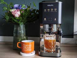 Krups Coffee Crush Experience Espresso-Vollautomat nebst Blumenwase und Dosen mit Tasse Kaffee und Glas mit Cold Brew