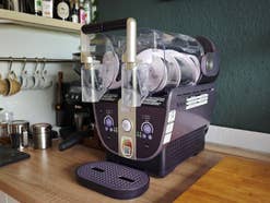 Eine Ninja Slushi Twist Slush-Maschine in Lila steht auf einer Küchentheke, nebst Schneidebrett, Kaffee-Utensilien etc.