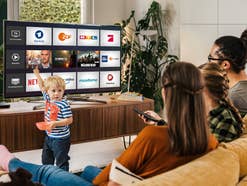 Eine Familie sitzt auf der Couch vor einem Fernseher, auf dem TV-Streaming-Dienste zu sehen sind.