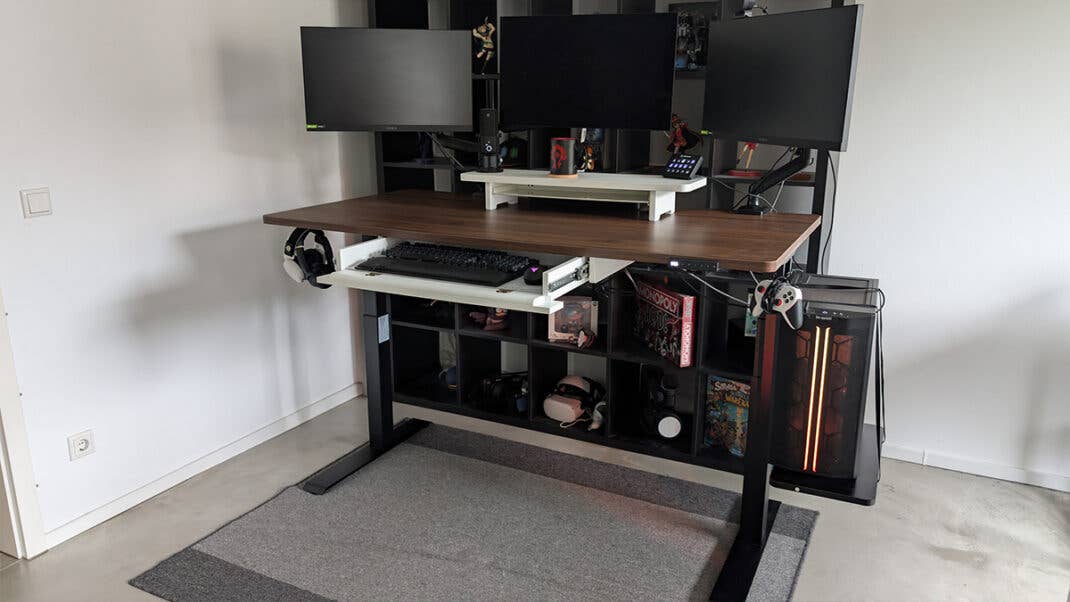 Drei Monitore auf einem höhenverstellbaren Schreibtisch mit einem Regal und Manga-Figuren im Hintergrund.