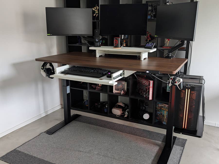 Drei Monitore auf einem höhenverstellbaren Schreibtisch mit einem Regal und Manga-Figuren im Hintergrund.