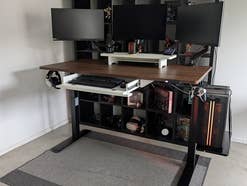 Drei Monitore auf einem höhenverstellbaren Schreibtisch mit einem Regal und Manga-Figuren im Hintergrund.