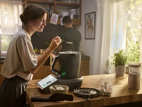 Eine Frau kocht in der Küche mit dem TM7 Thermomix von Vorwerk währen ihr Mann mit dem Rücken zu ihr steht.