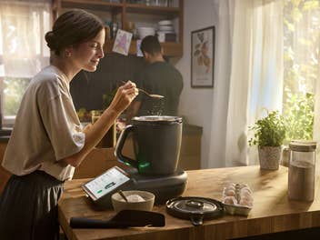 Eine Frau kocht in der Küche mit dem TM7 Thermomix von Vorwerk währen ihr Mann mit dem Rücken zu ihr steht.
