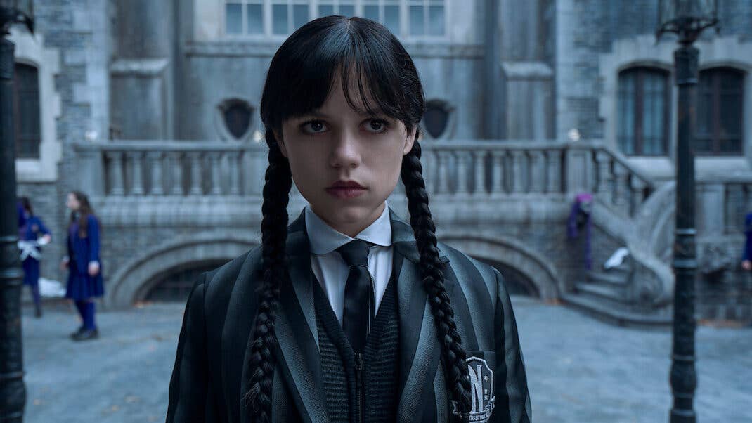 Wednesday Addams steht in einem dunklen Outfit vor einem gotischen Herrenhaus und sieht ernst aus.
