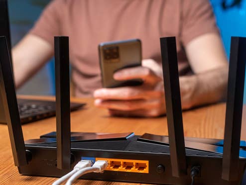 Warnung vor WLAN-Router-Verknappung Ein WLAN-Router im Vordergrund, im Hintergrund ein Mensch mit Laptop und Handy
