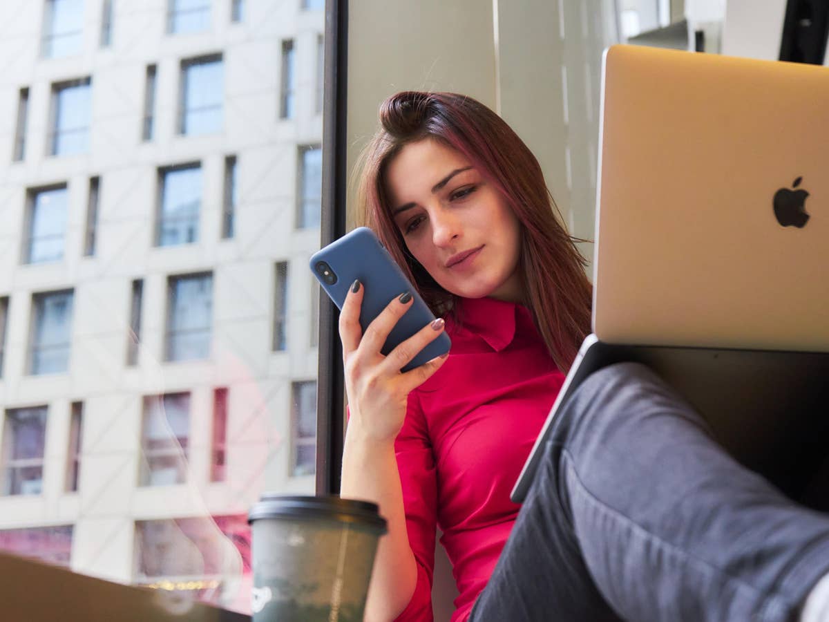 Eine Frau sitzt mit Laptop, Smartphone und einem Coffee to go auf einer Fensterbank