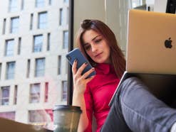 Eine Frau sitzt mit Laptop, Smartphone und einem Coffee to go auf einer Fensterbank